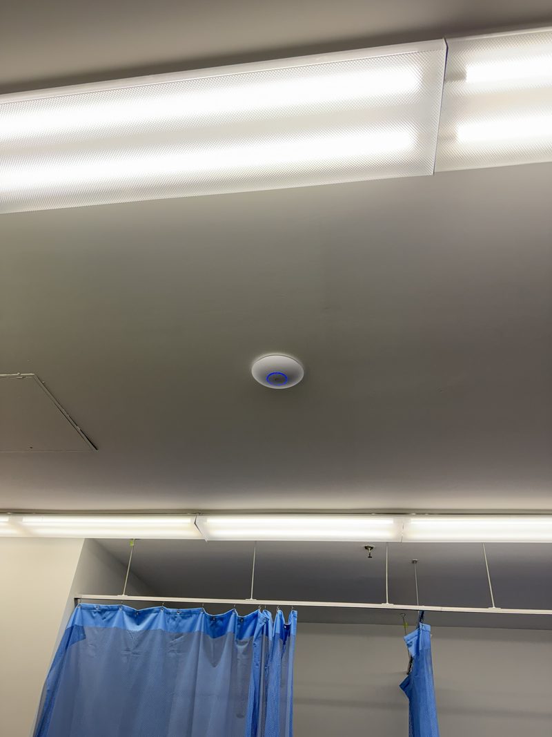 A white ceiling-mounted access point with a blue status ring, above a treatment area with blue privacy curtains.