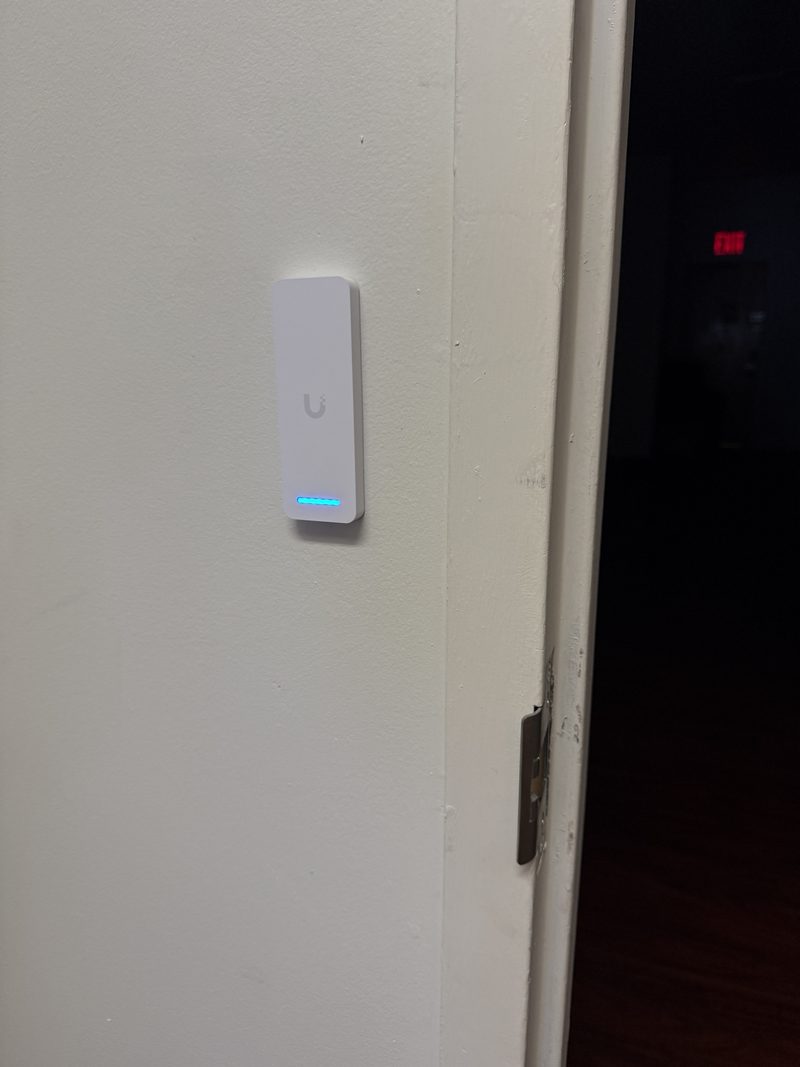 A slim white access-control reader with a blue light strip, mounted on a wall beside a door frame.