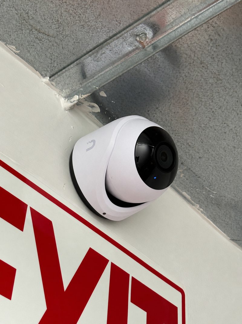 A white dome security camera mounted beside a red exit sign, under exposed steel.