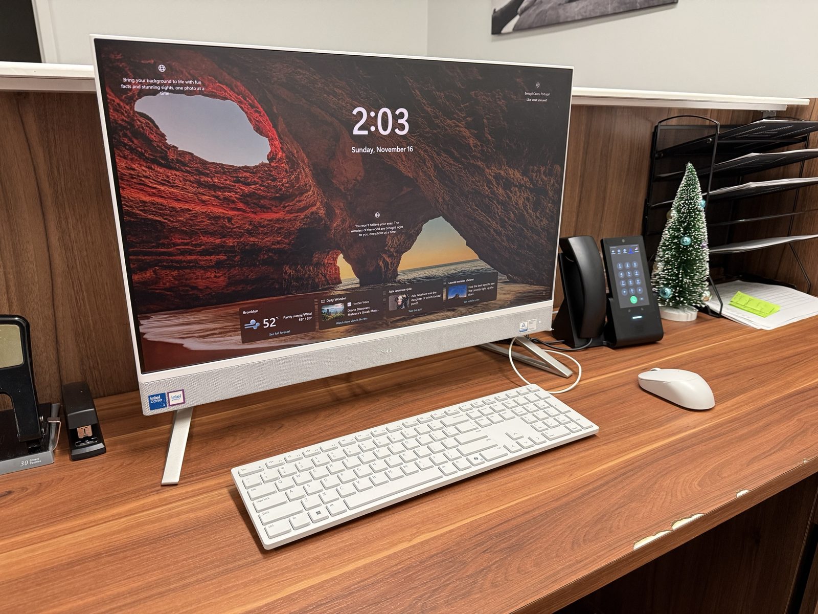 A white all-in-one desktop computer, keyboard and mouse on a wooden front desk, with a black touchscreen desk phone beside it.