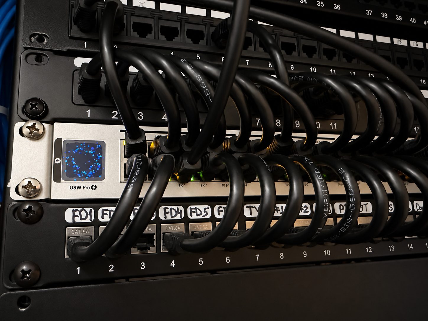 A network rack with a managed switch and two patch panels. Black patch cables loop from the switch to labeled ports; the front-desk runs are hand-labeled FD1 through FD5.